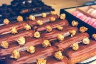 A selection of chocolate-covered eclairs topped with caramel popcorn is arranged neatly on a tray. The background shows a box containing pink macarons, adding a touch of color contrast.