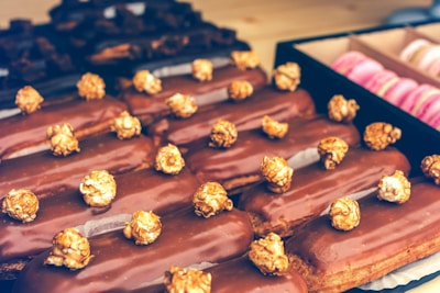 A selection of chocolate-covered eclairs topped with caramel popcorn is arranged neatly on a tray. The background shows a box containing pink macarons, adding a touch of color contrast.