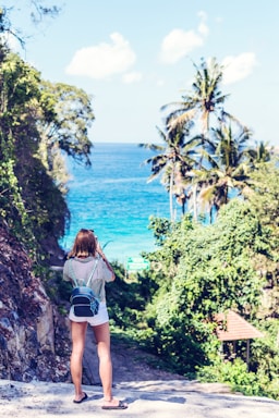 A woman stands on a sandy path in a tropical location, taking a photo of the vibrant blue ocean. She is wearing a striped shirt, white shorts, flip-flops, and a backpack. Tall palm trees and lush greenery surround the path, leading down to the water.