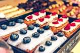 An assortment of pastries is displayed with eclairs topped with blueberries and others with raspberries. The eclairs have a smooth, creamy glaze, and are arranged neatly in a pastry box. The background shows a variety of other pastries with different toppings and textures.