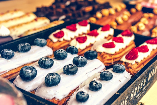 An assortment of pastries is displayed with eclairs topped with blueberries and others with raspberries. The eclairs have a smooth, creamy glaze, and are arranged neatly in a pastry box. The background shows a variety of other pastries with different toppings and textures.