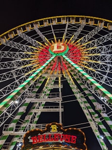 A brightly illuminated Ferris wheel during nighttime, showcasing vibrant green, red, and yellow lights. The wheel features a circular design with a large 'B' in the center and a sign reading 'BELLEVUE' at the bottom. The metal structure consists of intricate lattice work supporting the cabins and lighting.