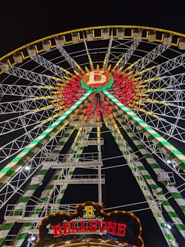 A brightly illuminated Ferris wheel during nighttime, showcasing vibrant green, red, and yellow lights. The wheel features a circular design with a large 'B' in the center and a sign reading 'BELLEVUE' at the bottom. The metal structure consists of intricate lattice work supporting the cabins and lighting.