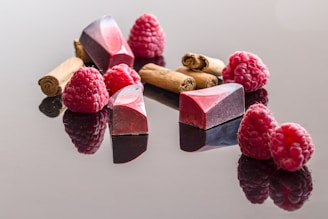 A collection of raspberries, cinnamon sticks, and elegantly crafted chocolates are arranged on a glossy surface. The chocolates have an angular, geometric design with a gradient of red and purple hues. The raspberries are fresh and vibrant, providing a rich contrast to the brown tones of the cinnamon sticks.