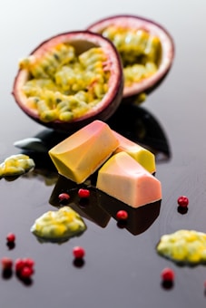 A halved passion fruit with its vibrant, yellow-green pulp is displayed alongside small, elegant pieces of gourmet chocolate. Red peppercorns are scattered around the arrangement, creating a visually appealing contrast against the glossy, dark surface.
