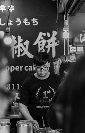 A person wearing glasses and a t-shirt reading '1st Taiwan Spirit' is standing at a counter. Bright lights hang from above, and there are other people partially visible around them. The backdrop features large Chinese characters and English words, including 'Pepper Cake'.