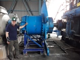 A man wearing a green hard hat and a wrist brace stands beside large industrial machinery in a factory setting. The machinery is primarily blue and consists of metal gears and components. The background shows a corrugated metal wall and other machinery.