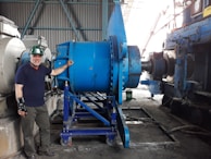 A man wearing a green hard hat and a wrist brace stands beside large industrial machinery in a factory setting. The machinery is primarily blue and consists of metal gears and components. The background shows a corrugated metal wall and other machinery.