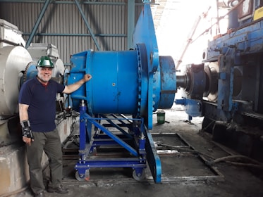 A man wearing a green hard hat and a wrist brace stands beside large industrial machinery in a factory setting. The machinery is primarily blue and consists of metal gears and components. The background shows a corrugated metal wall and other machinery.