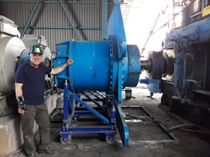 A man wearing a green hard hat and a wrist brace stands beside large industrial machinery in a factory setting. The machinery is primarily blue and consists of metal gears and components. The background shows a corrugated metal wall and other machinery.