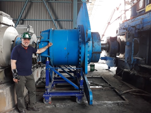 A man wearing a green hard hat and a wrist brace stands beside large industrial machinery in a factory setting. The machinery is primarily blue and consists of metal gears and components. The background shows a corrugated metal wall and other machinery.