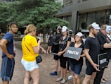 People are participating in a demonstration or protest. Most of them are wearing Guy Fawkes masks and black clothing. They are holding signs that read 'Truth'. Two individuals are engaged in a conversation on the left, one wearing a yellow shirt. The setting is an urban area with trees and buildings in the background.