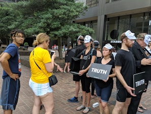 People are participating in a demonstration or protest. Most of them are wearing Guy Fawkes masks and black clothing. They are holding signs that read 'Truth'. Two individuals are engaged in a conversation on the left, one wearing a yellow shirt. The setting is an urban area with trees and buildings in the background.
