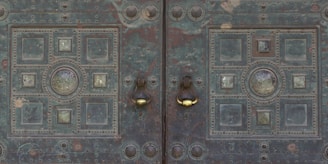A detailed, ornate metal door with symmetrical patterns featuring circular and square elements. The surface shows signs of aging with patina and wear, and there are two distinct metal door knockers with a polished, shiny appearance.
