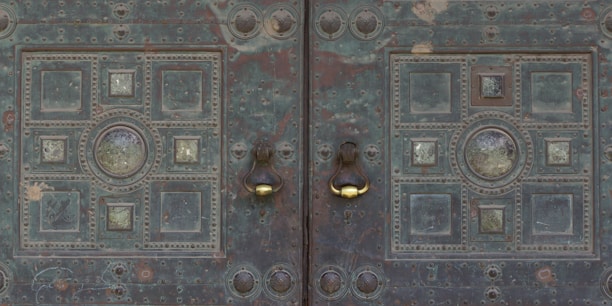 A detailed, ornate metal door with symmetrical patterns featuring circular and square elements. The surface shows signs of aging with patina and wear, and there are two distinct metal door knockers with a polished, shiny appearance.