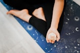 A person is sitting on a blue yoga mat adorned with celestial patterns, holding a crystal in their open hand, with bare feet visible in a relaxed pose.