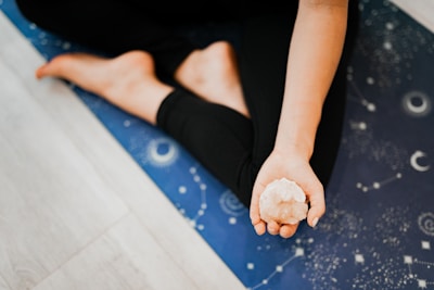 A person is sitting on a blue yoga mat adorned with celestial patterns, holding a crystal in their open hand, with bare feet visible in a relaxed pose.