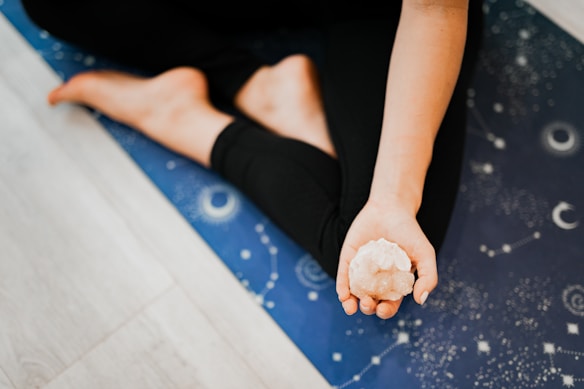 A person is sitting on a blue yoga mat adorned with celestial patterns, holding a crystal in their open hand, with bare feet visible in a relaxed pose.