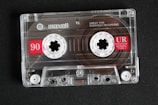 A transparent audio cassette tape with branding and labels displayed on a dark surface. The spools and magnetic tape are visible inside the clear plastic casing. Red labels with white text indicate the tape length and type.