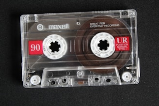 A transparent audio cassette tape with branding and labels displayed on a dark surface. The spools and magnetic tape are visible inside the clear plastic casing. Red labels with white text indicate the tape length and type.
