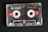 A transparent audio cassette tape with branding and labels displayed on a dark surface. The spools and magnetic tape are visible inside the clear plastic casing. Red labels with white text indicate the tape length and type.