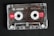 A transparent audio cassette tape with branding and labels displayed on a dark surface. The spools and magnetic tape are visible inside the clear plastic casing. Red labels with white text indicate the tape length and type.