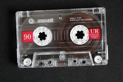 A transparent audio cassette tape with branding and labels displayed on a dark surface. The spools and magnetic tape are visible inside the clear plastic casing. Red labels with white text indicate the tape length and type.