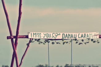 A sign made of wood and vines with the words 'I MISS YOU' followed by a heart drawing and 'DANAU CARAMIN' in large, painted letters. The sign is attached to wooden poles, set against a hazy sky and distant foliage.