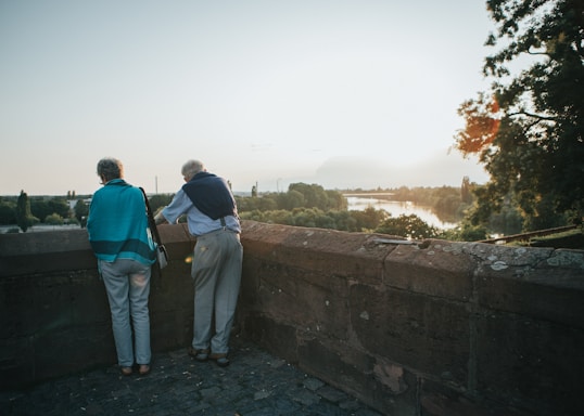Two older adults stand by a stone wall overlooking a scenic landscape with a river, surrounded by trees and bathed in the warm light of a setting or rising sun.
