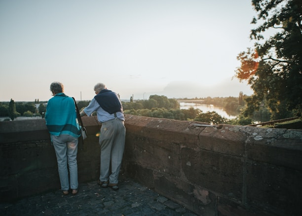 Two older adults stand by a stone wall overlooking a scenic landscape with a river, surrounded by trees and bathed in the warm light of a setting or rising sun.