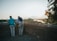 Two older adults stand by a stone wall overlooking a scenic landscape with a river, surrounded by trees and bathed in the warm light of a setting or rising sun.