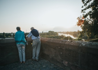 Two older adults stand by a stone wall overlooking a scenic landscape with a river, surrounded by trees and bathed in the warm light of a setting or rising sun.