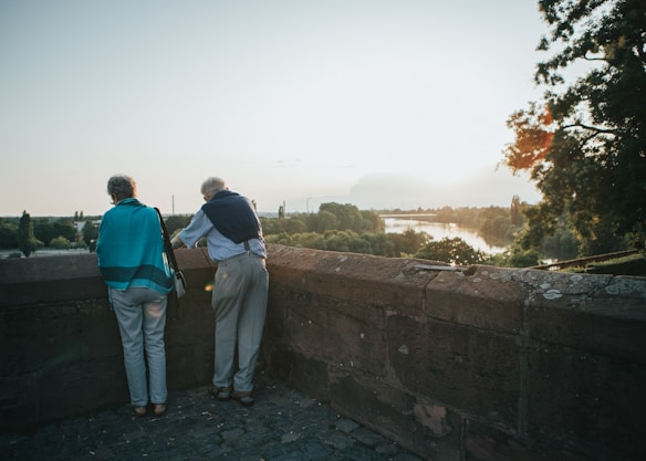 Two older adults stand by a stone wall overlooking a scenic landscape with a river, surrounded by trees and bathed in the warm light of a setting or rising sun.