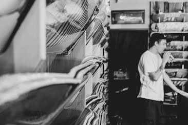 A black and white image featuring a narrow space filled with skateboards displayed on racks along both sides. A person wearing a white shirt is blurred in the background, possibly in motion or talking.