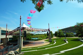 An outdoor amphitheater with a modern canopy structure and colorful flags strung across the top. The area is surrounded by well-maintained grass and pathways, with stone seating arranged in semi-circles. Trees and a body of water are visible in the background, along with a partly cloudy sky.