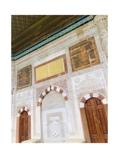 An ornate wall featuring intricate marble carvings, decorative arches, wooden doors, and embellished with gold and red accents. The top section of the wall is adorned with Arabic inscriptions and intricate tile-work in green and gold hues.