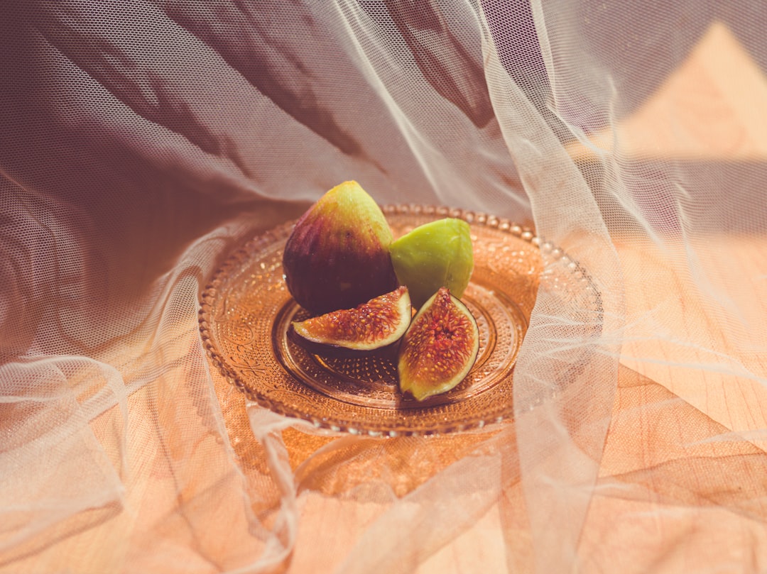 Several ripe figs served on an ornate glass plate behind wispy gauze fabric drapery drenched in sunlight coming through the window.