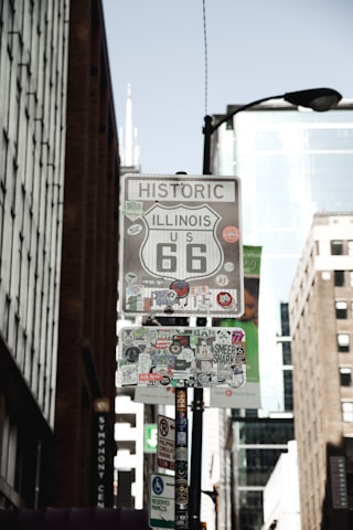 A street sign for Historic US Route 66 in Illinois is covered with numerous stickers. The background shows tall buildings with glass and brick facades, indicating an urban environment.