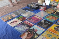 A collection of vintage vinyl records is displayed on a blue cloth. Various album covers with colorful artwork and text are visible, reflecting a range of music genres. There are also some cassette tapes and other miscellaneous items on the table. The setting appears to be an outdoor market, with a tiled surface visible underneath.