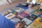 A collection of vintage vinyl records is displayed on a blue cloth. Various album covers with colorful artwork and text are visible, reflecting a range of music genres. There are also some cassette tapes and other miscellaneous items on the table. The setting appears to be an outdoor market, with a tiled surface visible underneath.