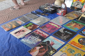 A collection of vintage vinyl records is displayed on a blue cloth. Various album covers with colorful artwork and text are visible, reflecting a range of music genres. There are also some cassette tapes and other miscellaneous items on the table. The setting appears to be an outdoor market, with a tiled surface visible underneath.