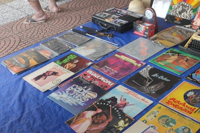 A collection of vintage vinyl records is displayed on a blue cloth. Various album covers with colorful artwork and text are visible, reflecting a range of music genres. There are also some cassette tapes and other miscellaneous items on the table. The setting appears to be an outdoor market, with a tiled surface visible underneath.