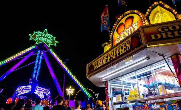 A lively amusement park scene at night featuring a brightly lit ride with green and yellow neon star decorations. A candy stand offering caramel apples and other treats is in the foreground. Crowds of people are gathered around enjoying the attractions.