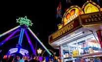 A lively amusement park scene at night featuring a brightly lit ride with green and yellow neon star decorations. A candy stand offering caramel apples and other treats is in the foreground. Crowds of people are gathered around enjoying the attractions.