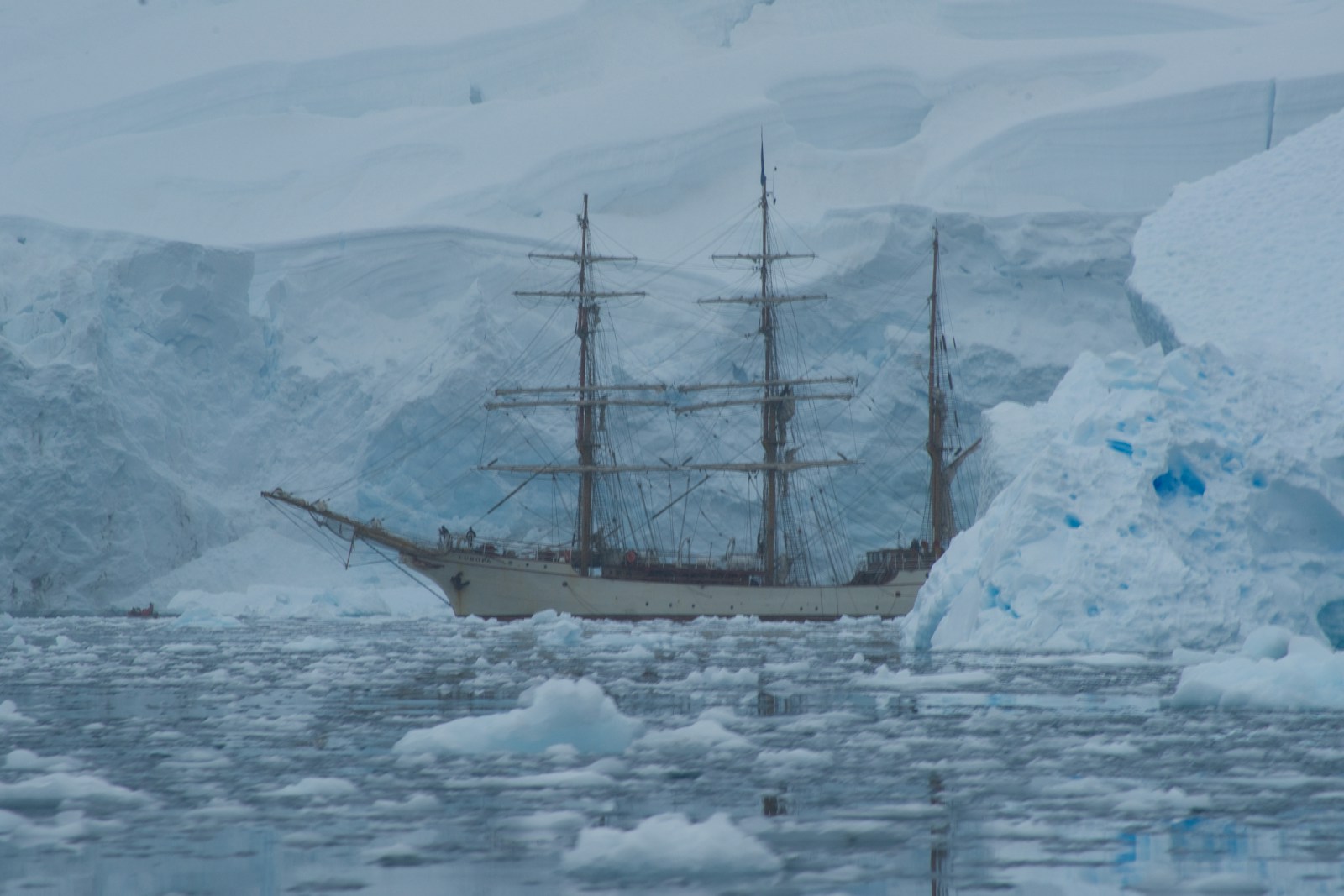 A three-masted sailing ship at anchor before an ice cliff, surrounded by floating ice in grey water