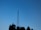 A tall metal radio or television tower stands against a clear blue sky, with silhouetted trees at the base.