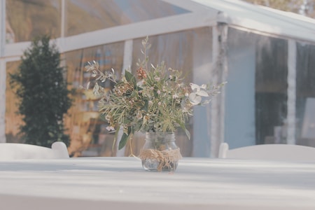 A rustic floral arrangement sits in a glass jar on a round white table. The jar is decorated with a twine bow and filled with a variety of greenery and small white flowers. In the background, a clear tent structure with sunlight filtering through can be seen along with some blurred foliage.