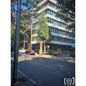 A multi-story modern office building is surrounded by trees. Several cars are parked along the street, and a row of motorcycles is lined up on the sidewalk. There is a no-entry traffic sign near a tree, and a lamppost is visible in the foreground.