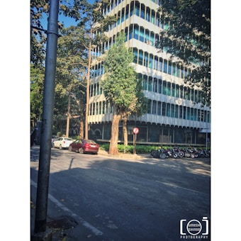 A multi-story modern office building is surrounded by trees. Several cars are parked along the street, and a row of motorcycles is lined up on the sidewalk. There is a no-entry traffic sign near a tree, and a lamppost is visible in the foreground.