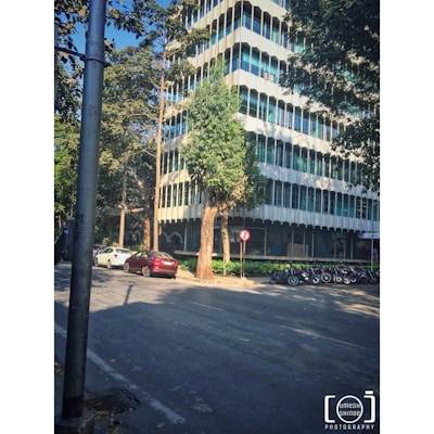 A multi-story modern office building is surrounded by trees. Several cars are parked along the street, and a row of motorcycles is lined up on the sidewalk. There is a no-entry traffic sign near a tree, and a lamppost is visible in the foreground.